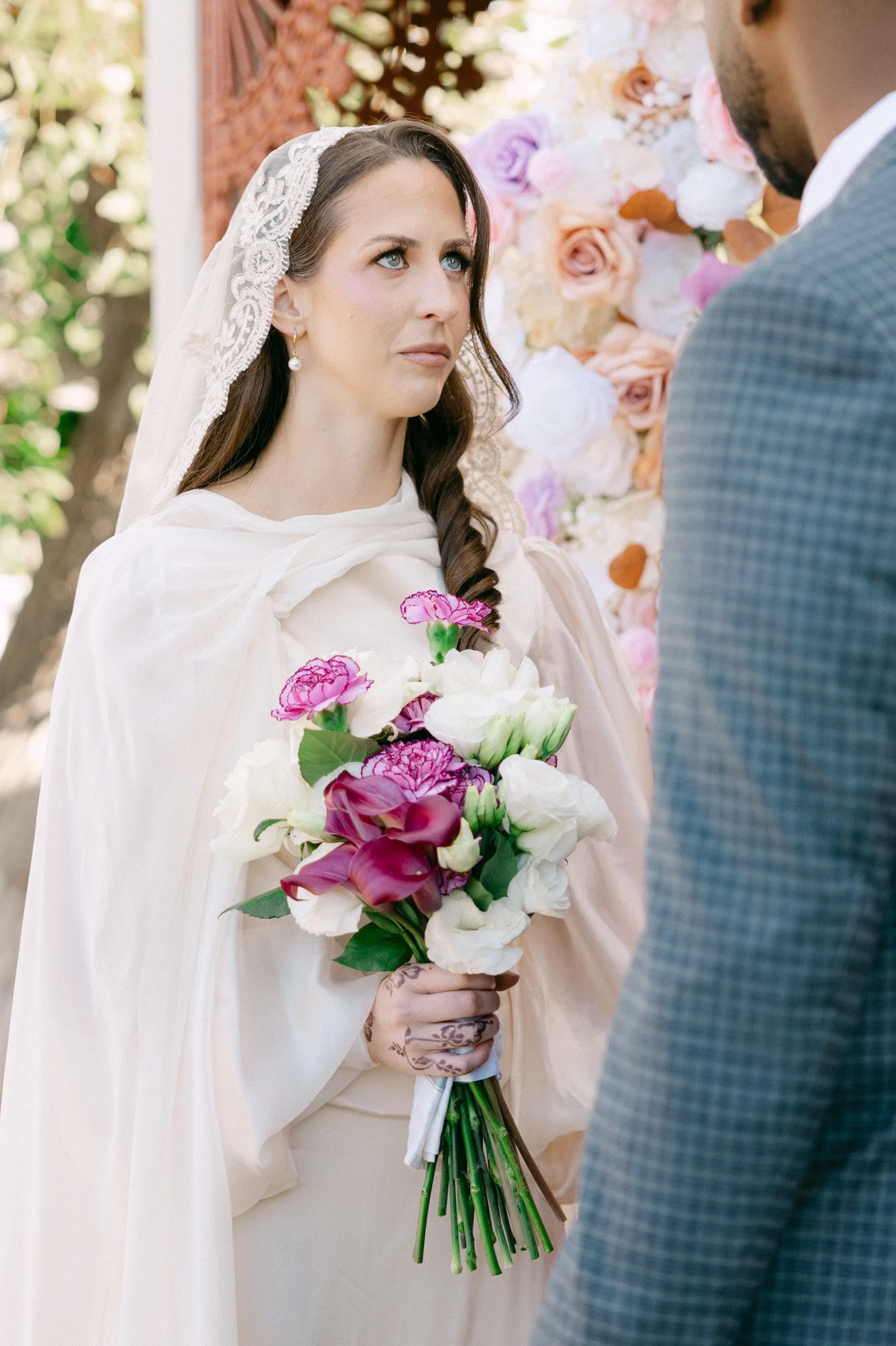 A woman at her wedding holding a bouquet of pink, white, and purple flowers, with a lace veil on her head and a white dress, standing in front of a floral backdrop, gazing at a man in a gray checkered suit.