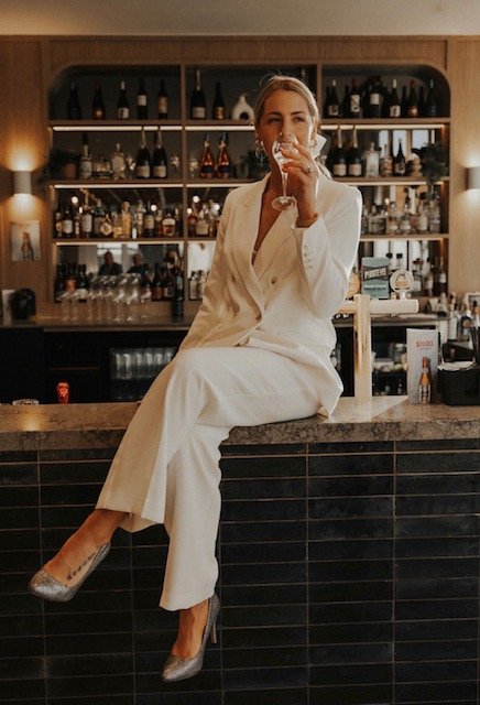 A woman in a white suit sitting on a bar counter, drinking from a glass, with a background of shelves filled with bottles of alcohol in a bar setting.