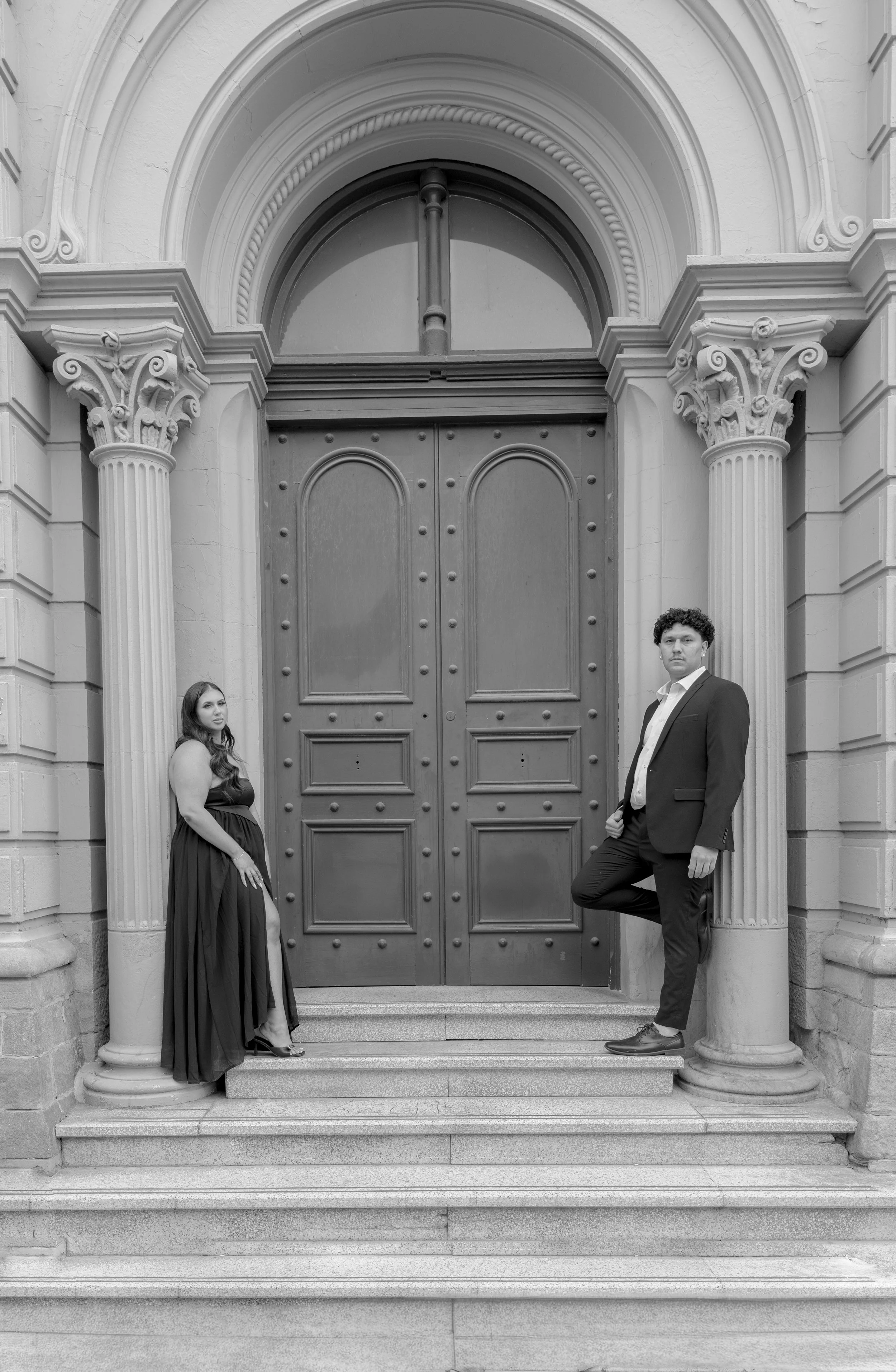 A black-and-white photograph of a man and a woman posed on the steps in front of a large wooden door with ornate columns on either side. The man is dressed in a suit and leaning against one of the columns, while the woman is wearing a strapless gown and standing near the other column.
