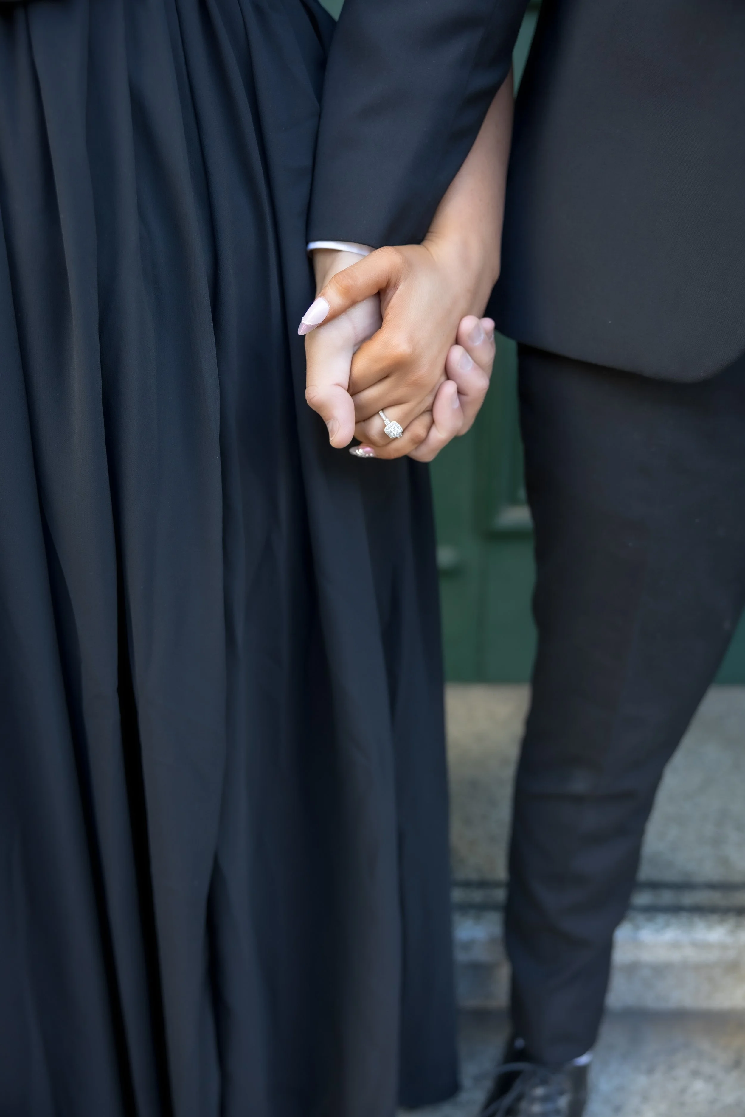 Close-up of a couple holding hands, with the woman wearing a graduation gown and ring, and the man in a suit.