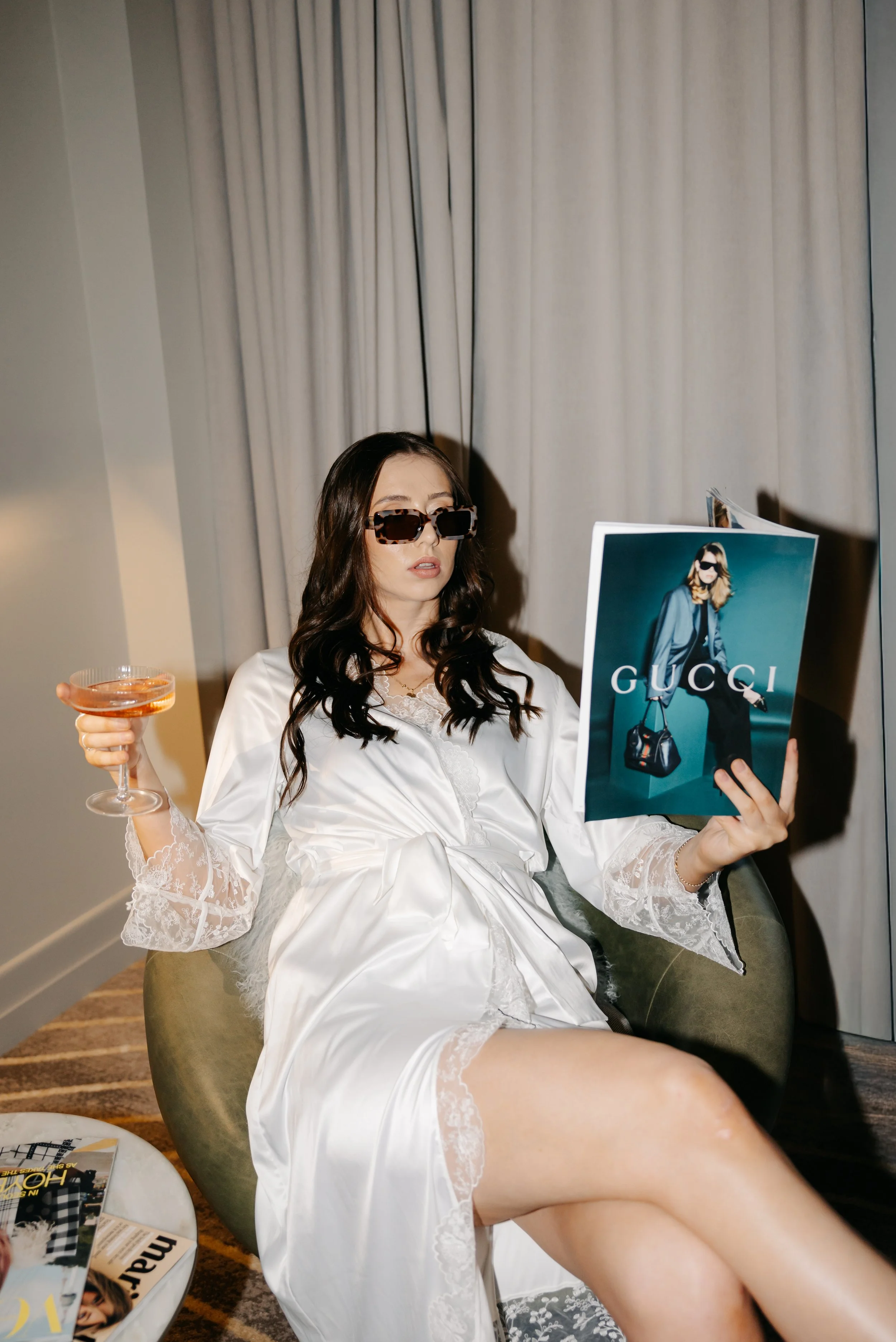 A woman wearing a satin white robe and dark sunglasses is sitting in a green chair holding a glass of pink wine. She is reading a Gucci fashion magazine and has long dark hair. There are curtains in the background and a small table with magazines in 