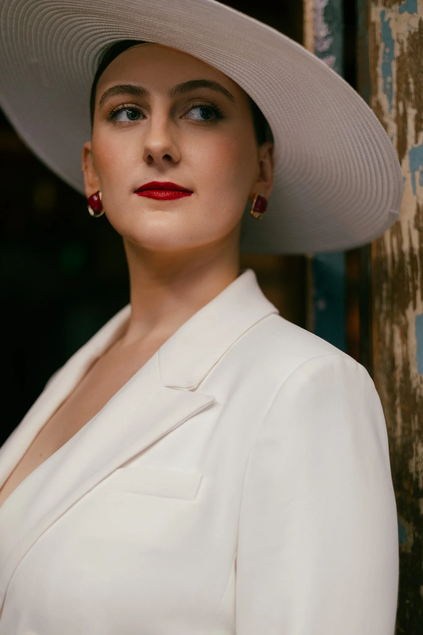A woman wearing a large white hat with a wide brim, a white blazer, red earrings, with makeup including red lipstick and eye makeup, standing near a rustic wooden wall.