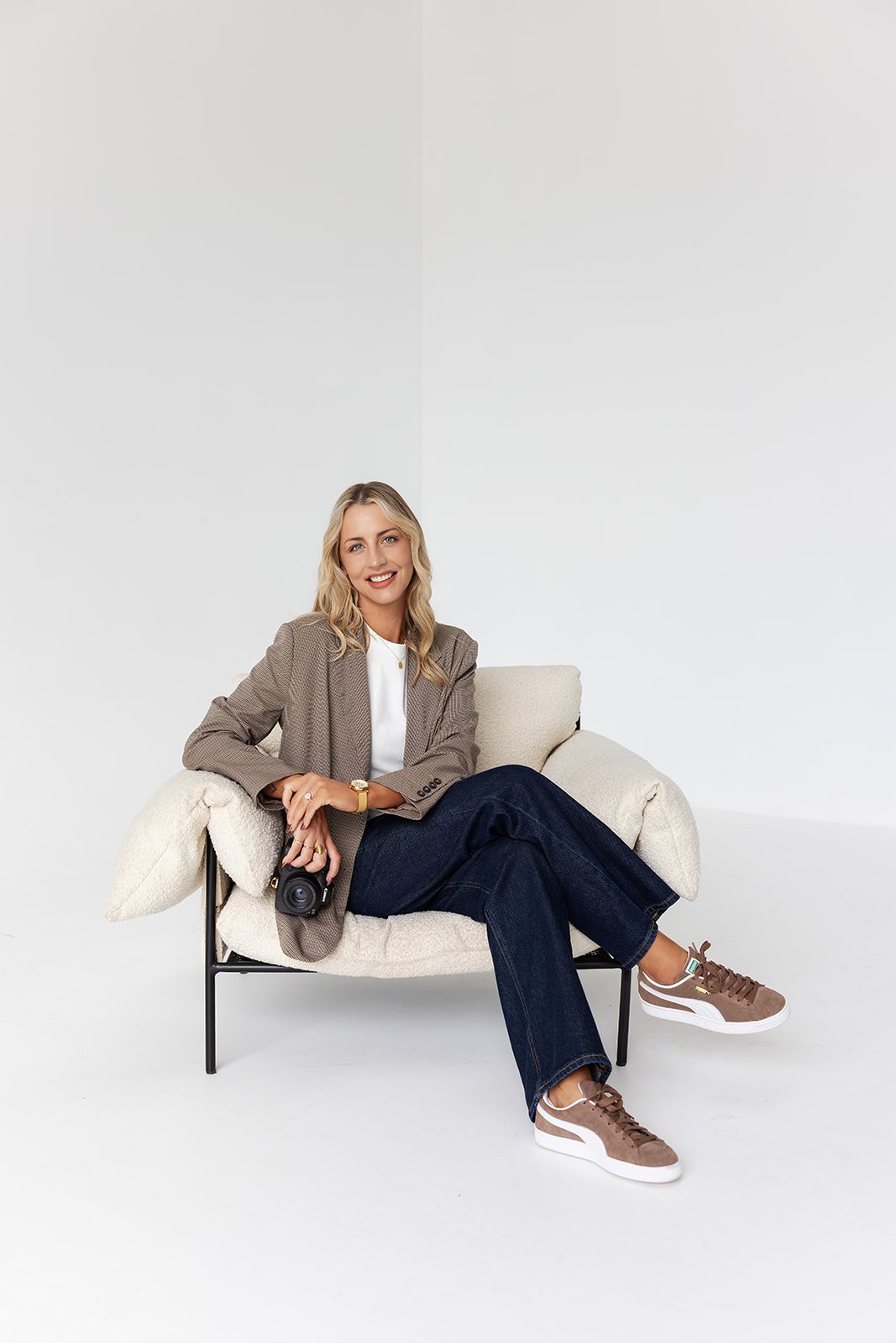 A woman sitting on a modern sofa in a minimalist white room, holding a camera and smiling.