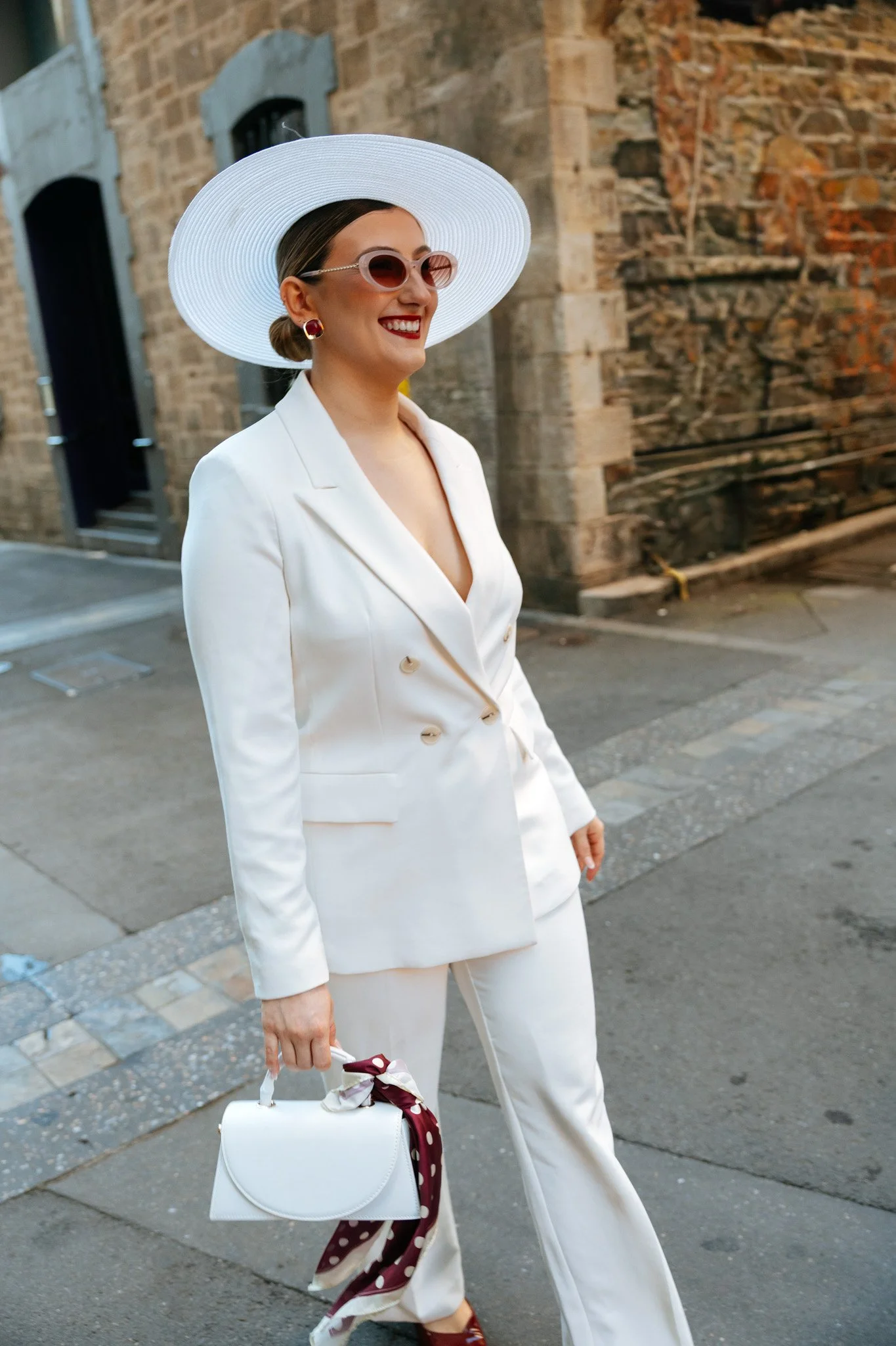 A woman wearing a white suit, large white hat, pink sunglasses, and red lipstick, smiles while walking on a street with brick buildings in the background. She carries a white handbag with a polka dot scarf tied to it.