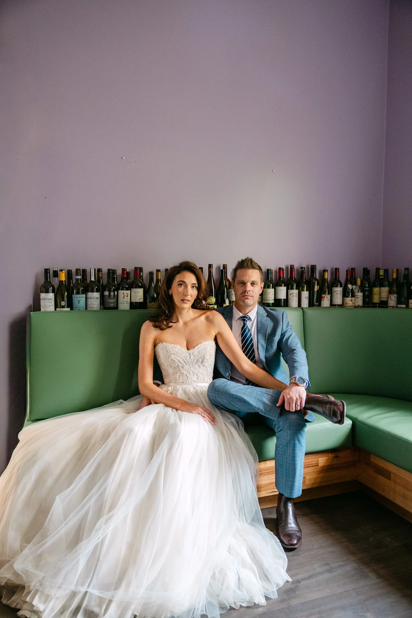 A bride and groom sitting on a green bench against a purple wall with wine bottles on a shelf behind them. The bride wears a strapless wedding dress, and the groom wears a blue suit with a tie and brown boots.