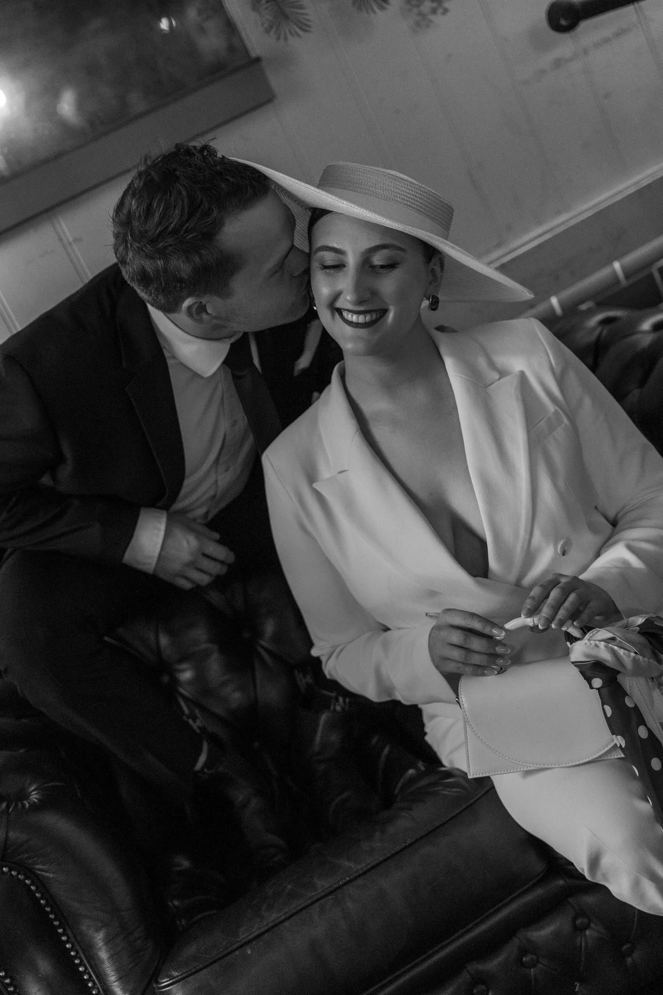 A black and white photograph of a man and a woman sitting on a leather couch. The man is kissing the woman on the cheek. The woman is smiling with her eyes closed, wearing a wide-brimmed hat and a white blazer. She is holding a cigarette in her right