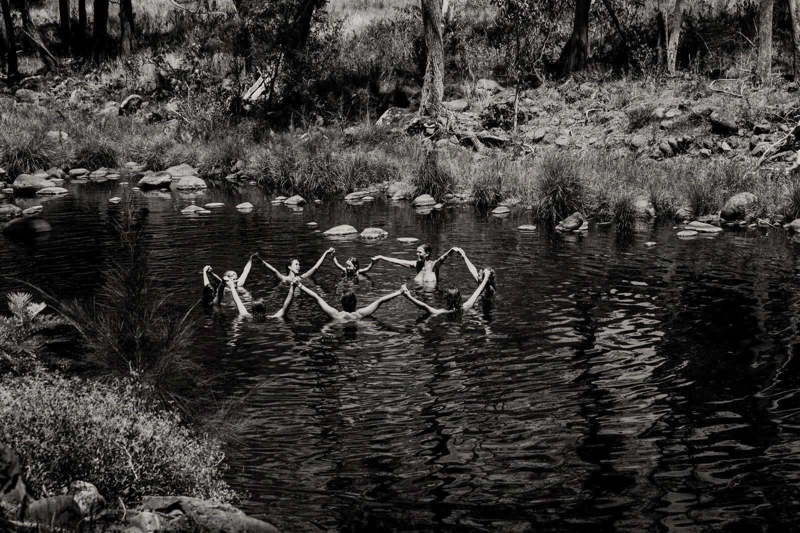 A group of people swimming in a river, holding hands in a circle, surrounded by trees and rocks.