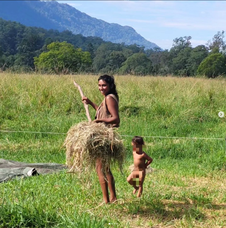 A smiling woman holding a stick and a bunch of hay standing next to a small child in a grassy field with mountains and trees in the background.