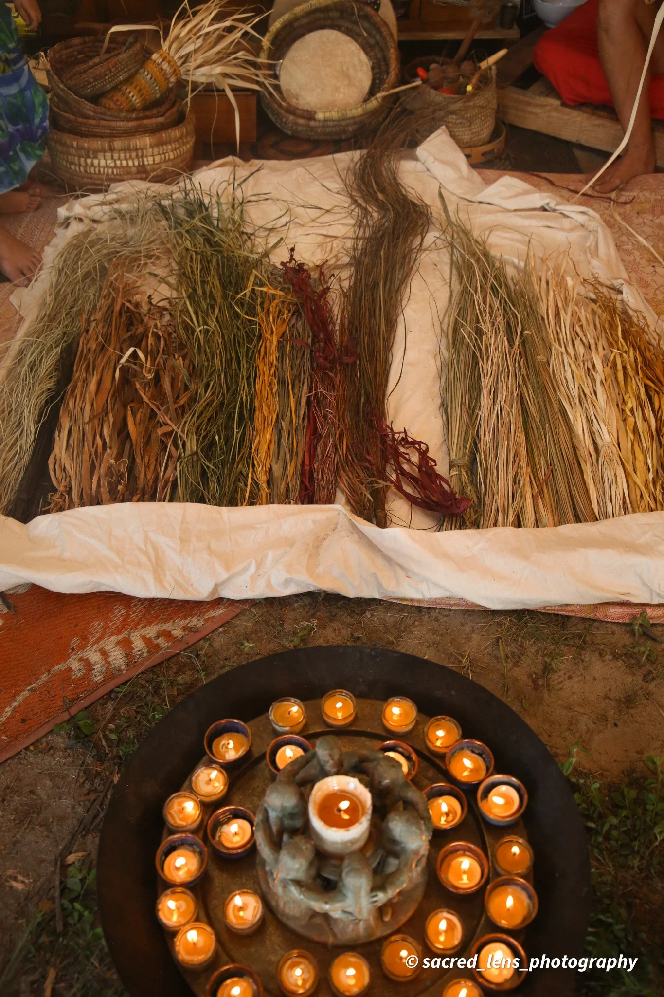 A circular arrangement of small lit candles surrounds a central decorative object made of shells. In the background, there are woven baskets filled with natural materials such as dried grasses and a drum, along with fabric and other traditional items, suggesting a cultural or spiritual setting.