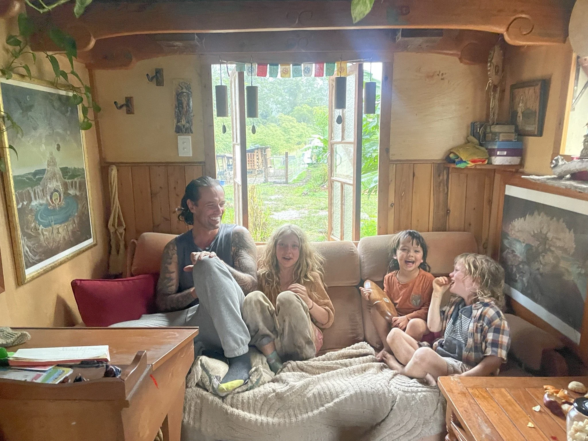 A group of smiling children and a man sitting on a couch inside a cozy wooden room with open windows showing green trees outside. The room is decorated with colorful flags, paintings, and various decorative items.