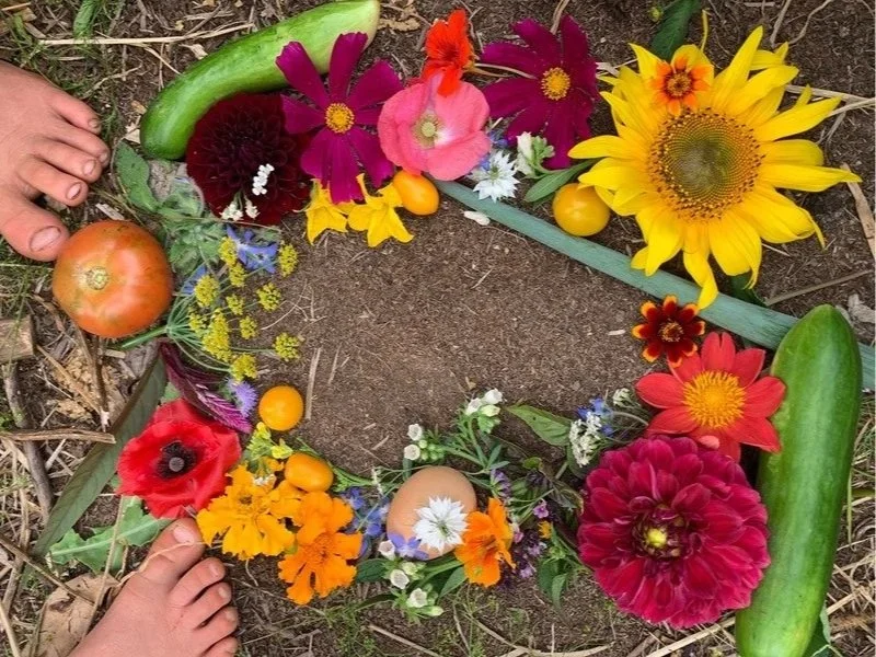 Colorful flower wreath on soil with tomatoes, egg, green cucumber, and fern leaves, and children's feet nearby.
