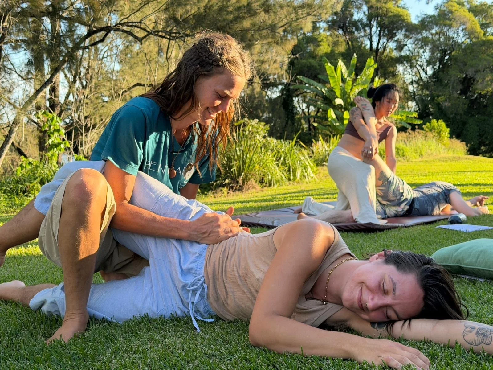 Women practicing yoga outdoors on grass with trees in the background, one woman lying on her side and another woman giving a massage, while two others are in the background practicing yoga poses.