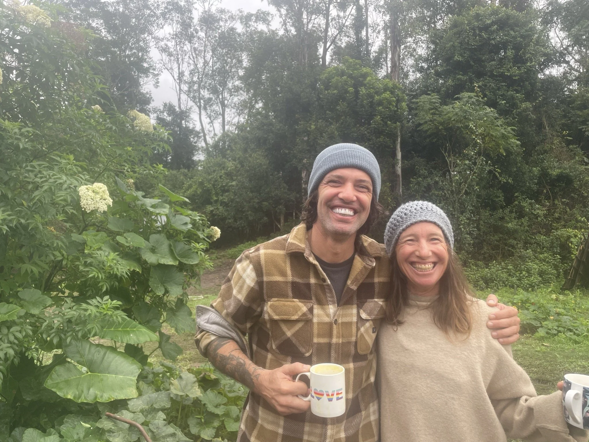 Two smiling people, a man and a woman, standing outdoors in a lush green garden, both wearing gray knit hats, with the man holding a mug with the word "LOVE" written on it.