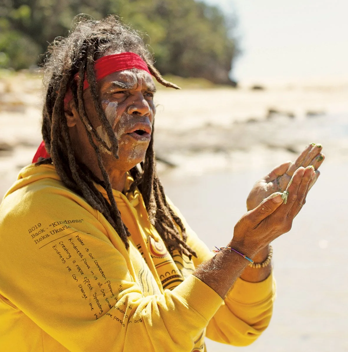 A man with dreadlocks, wearing a red headband and a yellow hoodie, stands on a beach with his hands raised and eyes closed, engaged in a spiritual practice.