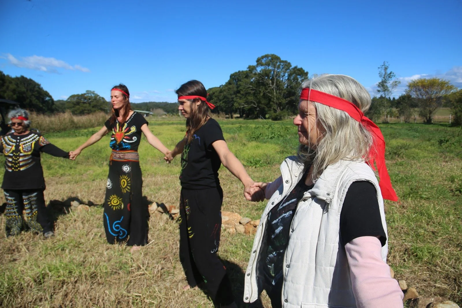 A group of diverse women holding hands in a circle outdoors on a sunny day, some wearing traditional Indigenous attire and others in casual clothing, with greenery and trees in the background.