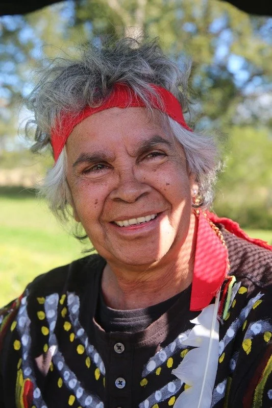Smiling elderly woman with gray curly hair, red headband, and colorful patterned shirt outdoors on a sunny day.