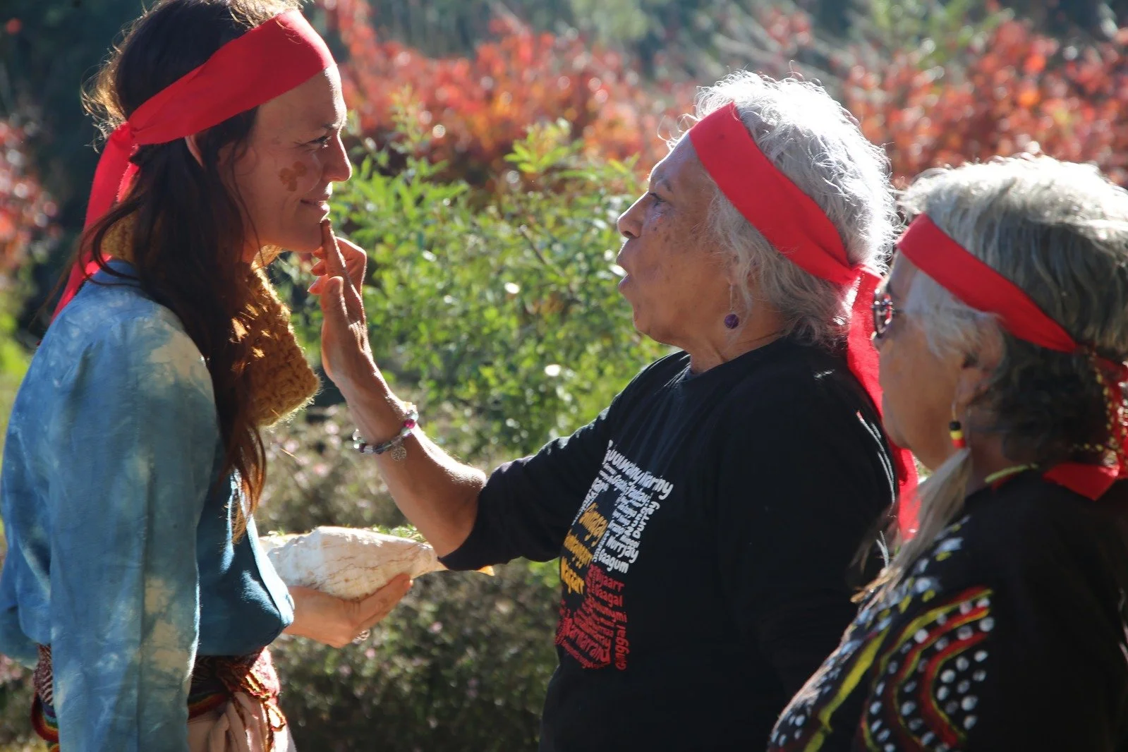 Three women in an outdoor setting wearing red headbands, with one woman touching another woman's face in a gentle gesture, while the third woman looks on. The women appear to be participating in a cultural or spiritual activity, with colorful foliage in the background.