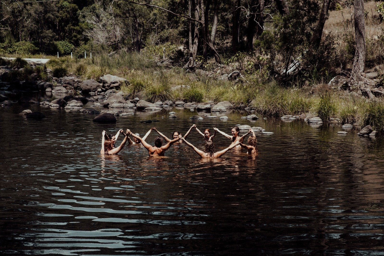 A group of people are forming a circle in a river, holding hands, surrounded by nature with trees and rocks along the riverbank.