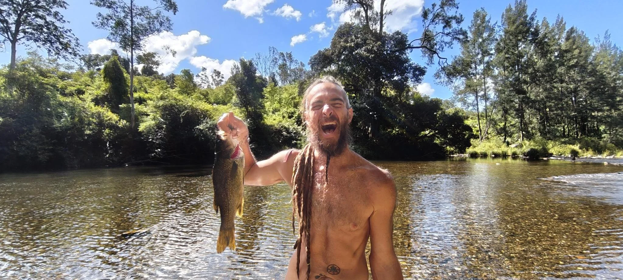 A man with long dreadlocks and a beard standing in a river, holding a fish in his right hand, and smiling widely. The background features lush green trees and a partly cloudy sky.