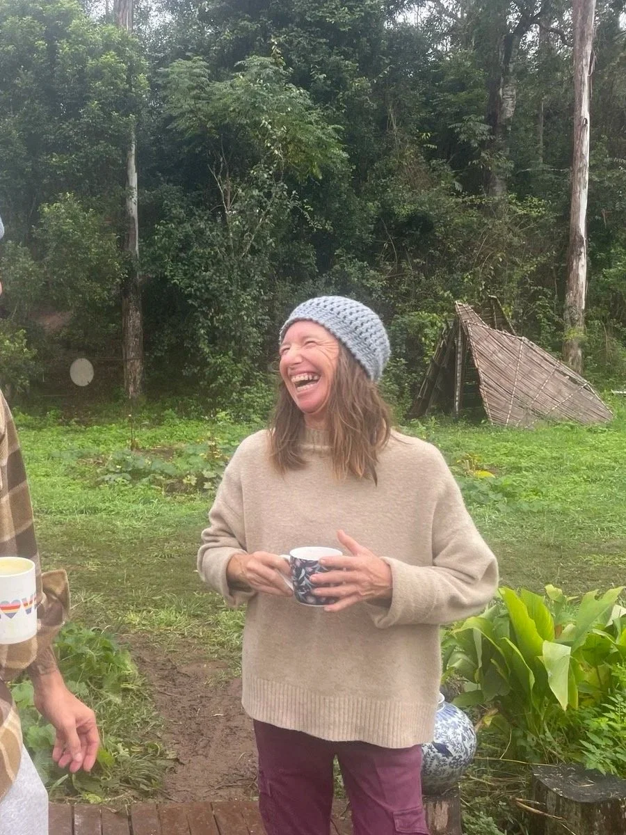 A woman laughing and holding a mug outdoors in a green, wooded area.