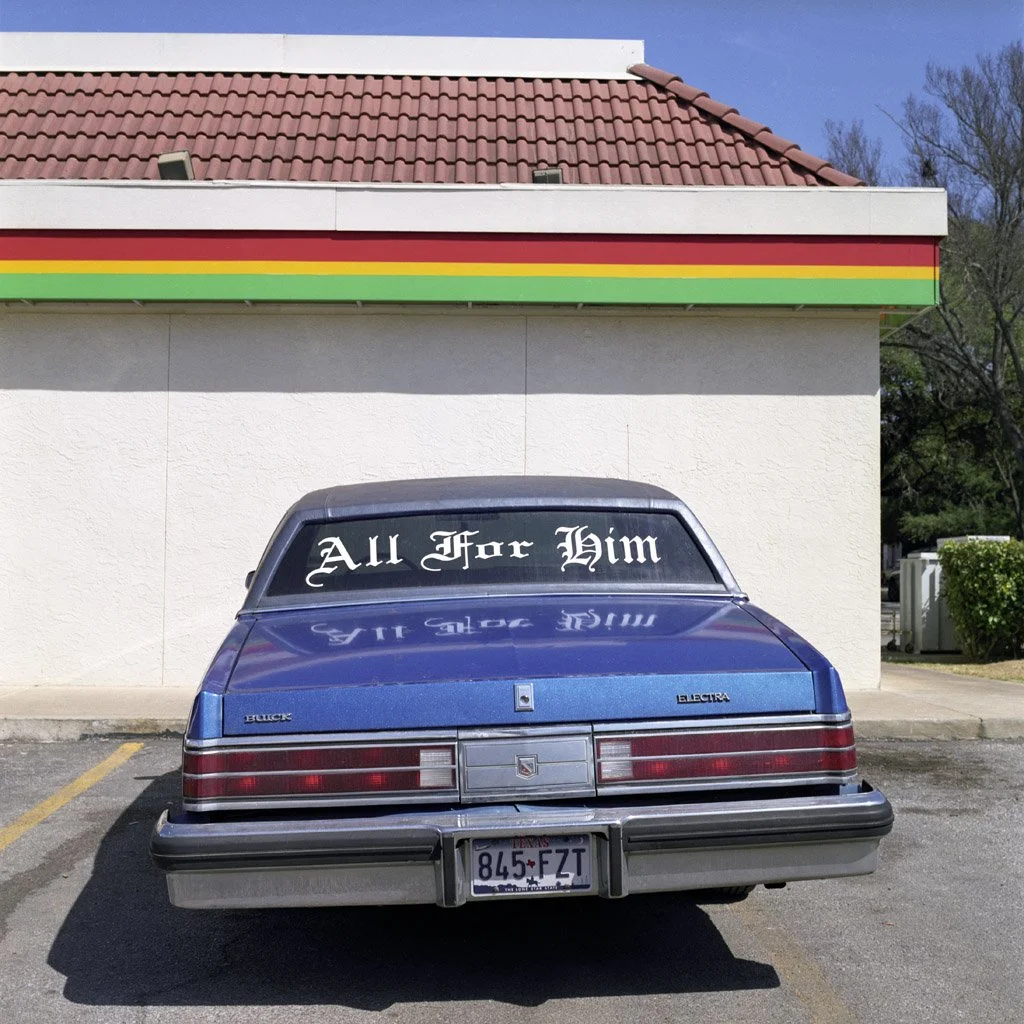 Blue Buick Electra car parked in front of a convenience store with a sign that says 'All For Him' on the rear window.