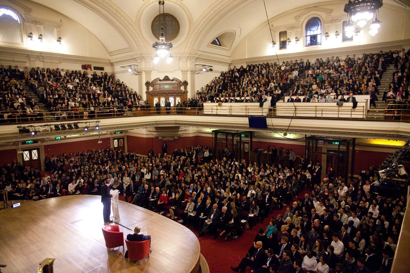 A large auditorium packed with people attending a formal event. A speaker stands at a podium on the stage, addressing the audience.