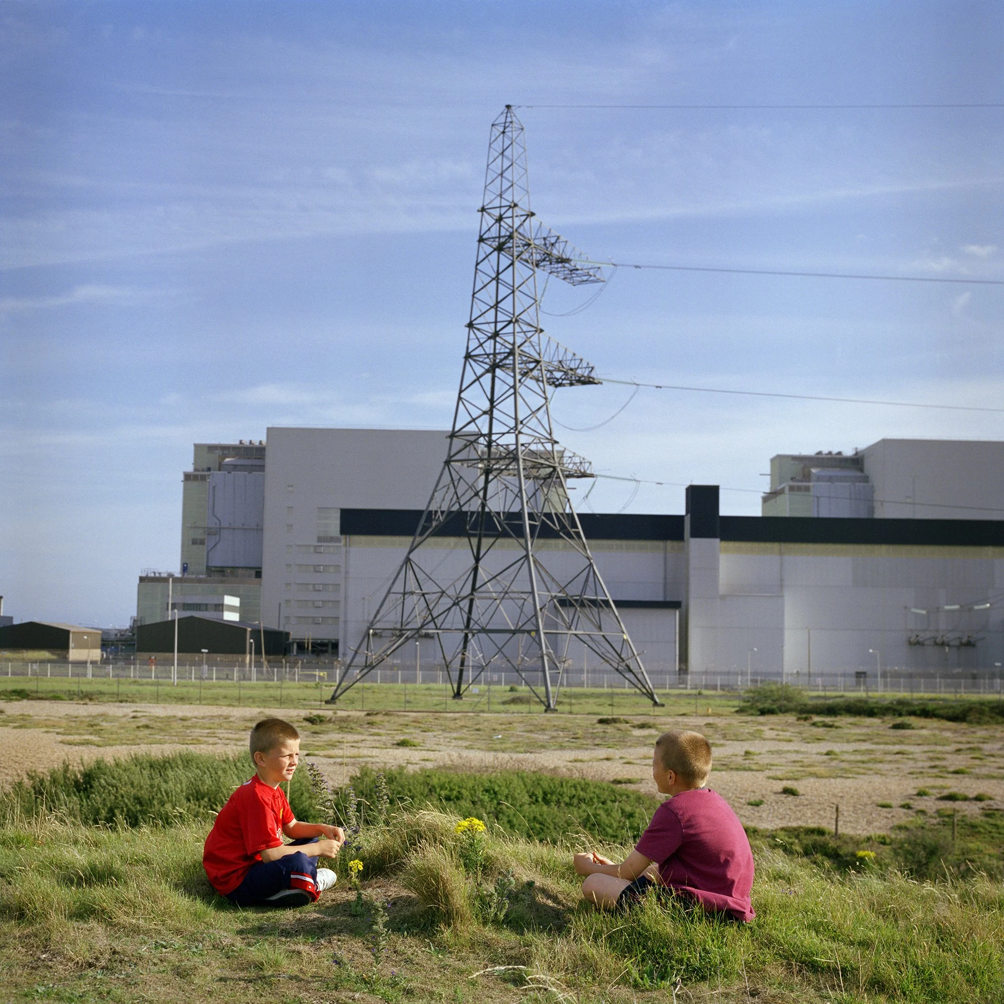 Two children sitting on grass in a field with flowers, engaged in conversation, with a large electrical transmission tower and modern buildings in the background under a partly cloudy sky.