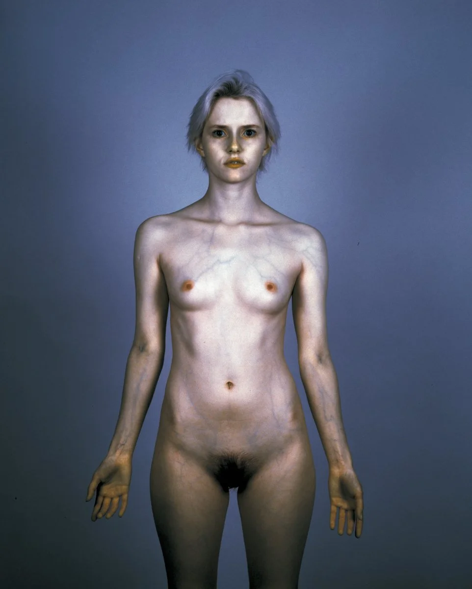 A digitally edited photograph of a woman with pale skin, short light-colored hair, and an expressionless face, standing against a plain background. Her body appears unnaturally elongated and thin, with exaggerated features.