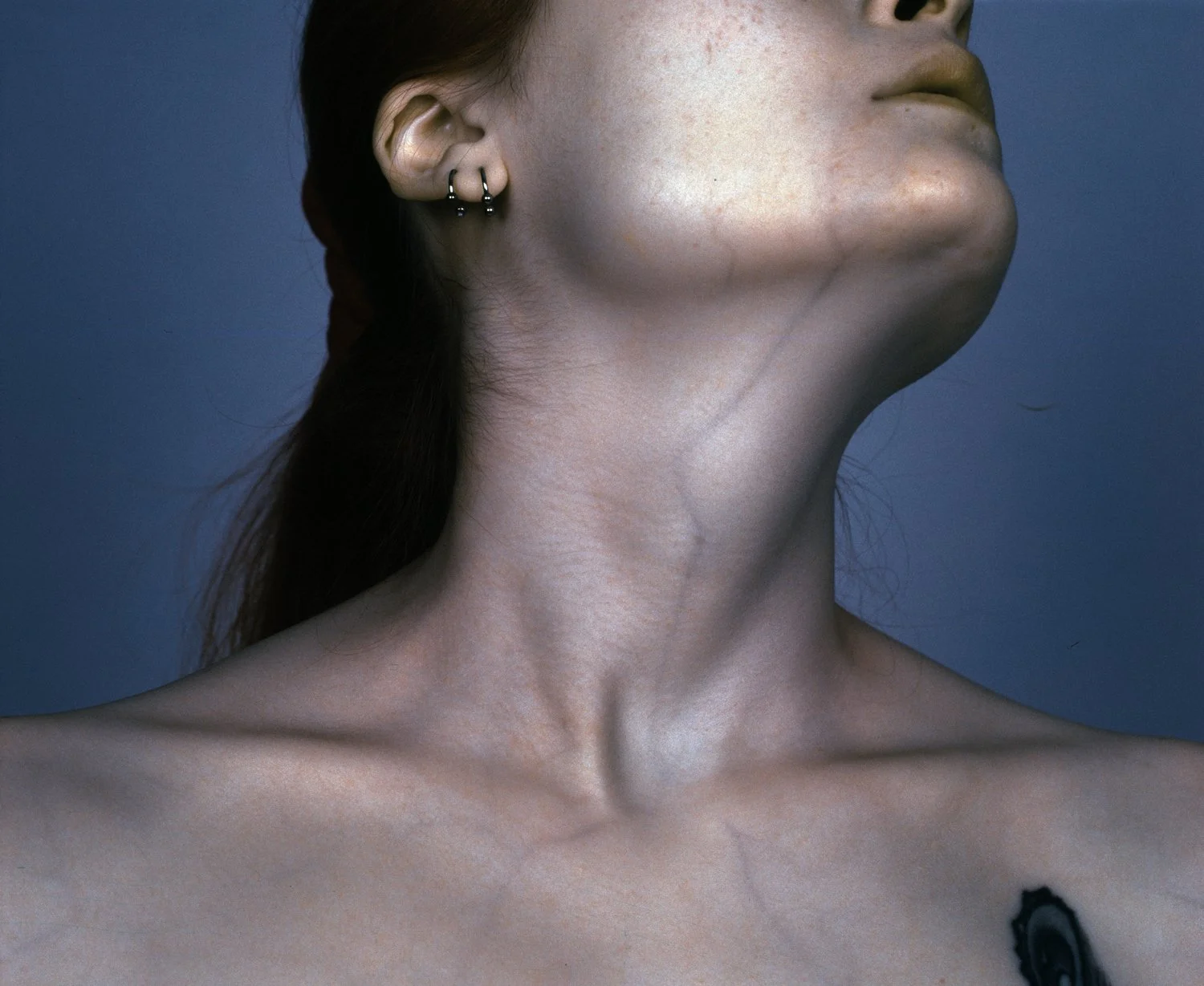Close-up of a woman's face and neck, with her head tilted back, showing her profile and shoulder, against a plain blue background.