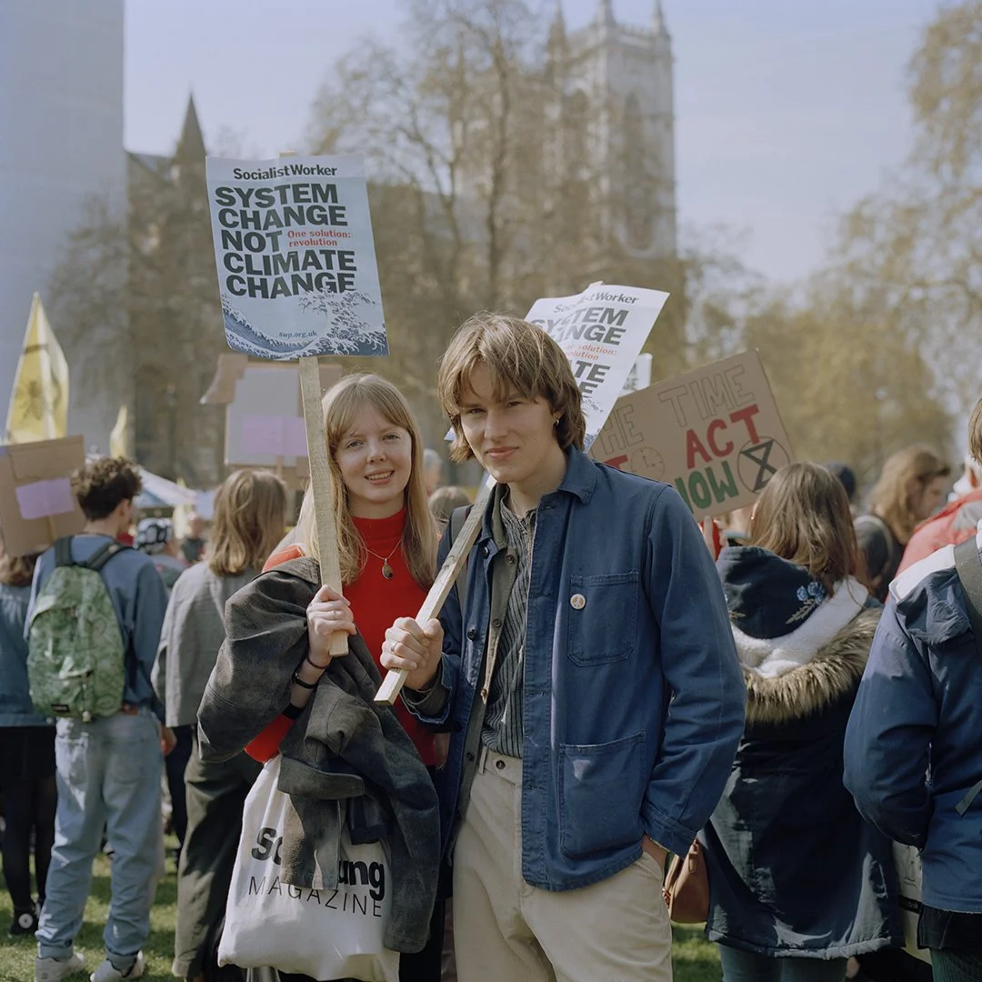 Two young people at a protest holding signs and surrounded by a crowd of protesters, with trees and a building in the background.