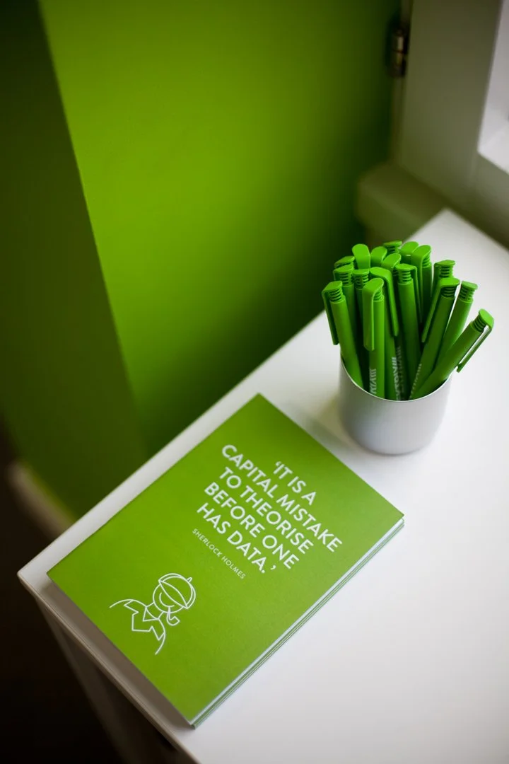Green brochures on a white table with a container of green pens. The brochure reads, "It is a capital mistake to theorise before one has data. Sherlock Holmes."