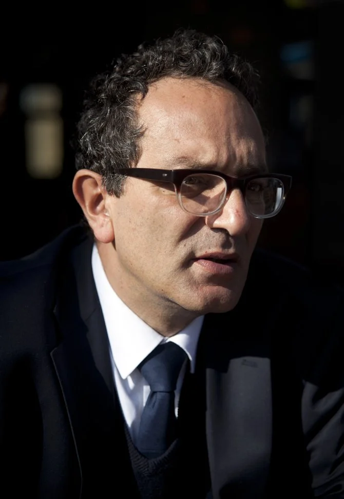 A man with curly dark hair and glasses, dressed in a suit and tie, outdoors with sunlight on his face.