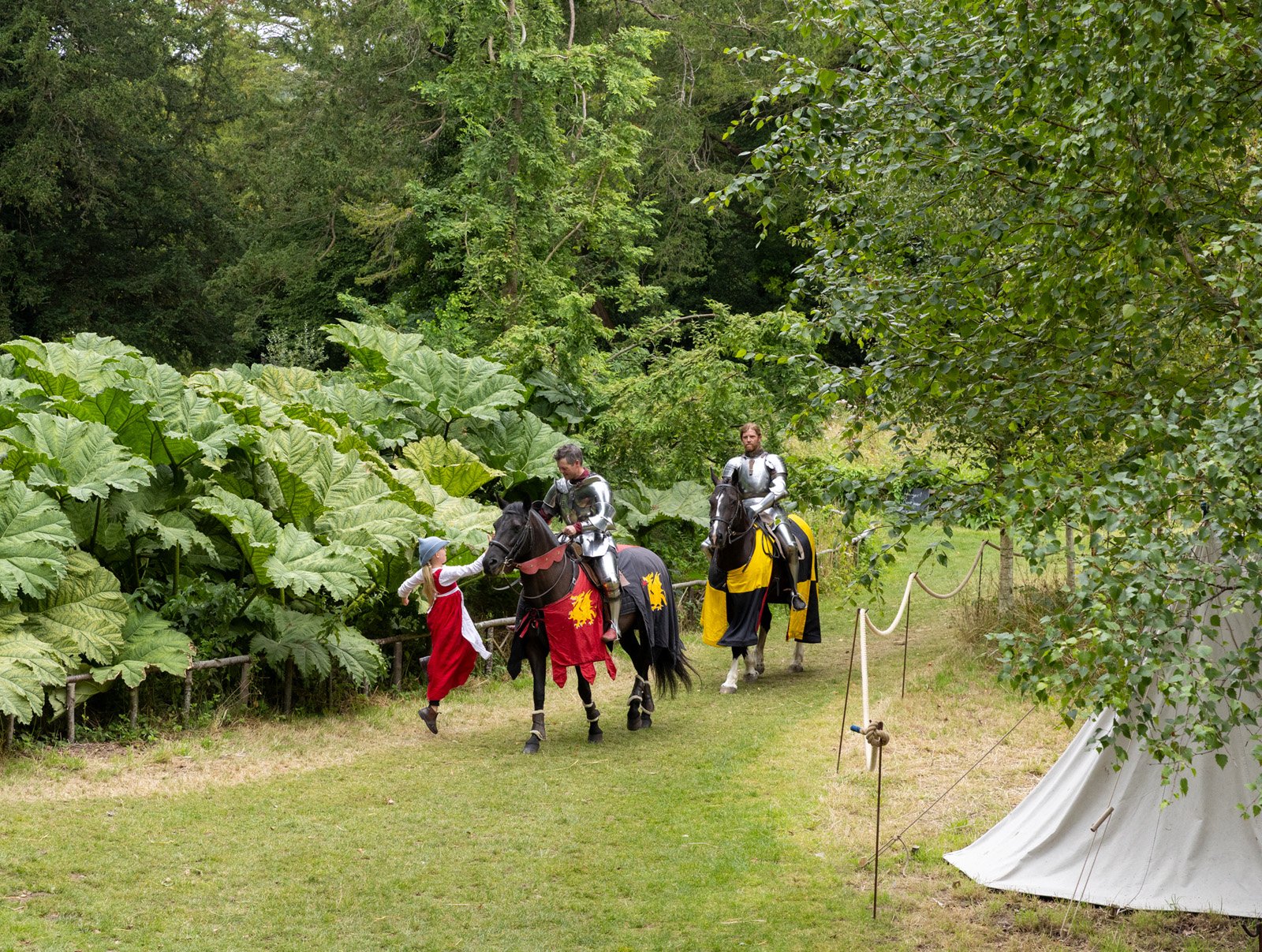 Two men dressed as medieval knights riding black horses in a lush green outdoor setting, interacting with a young girl in a red dress and blue hat.
