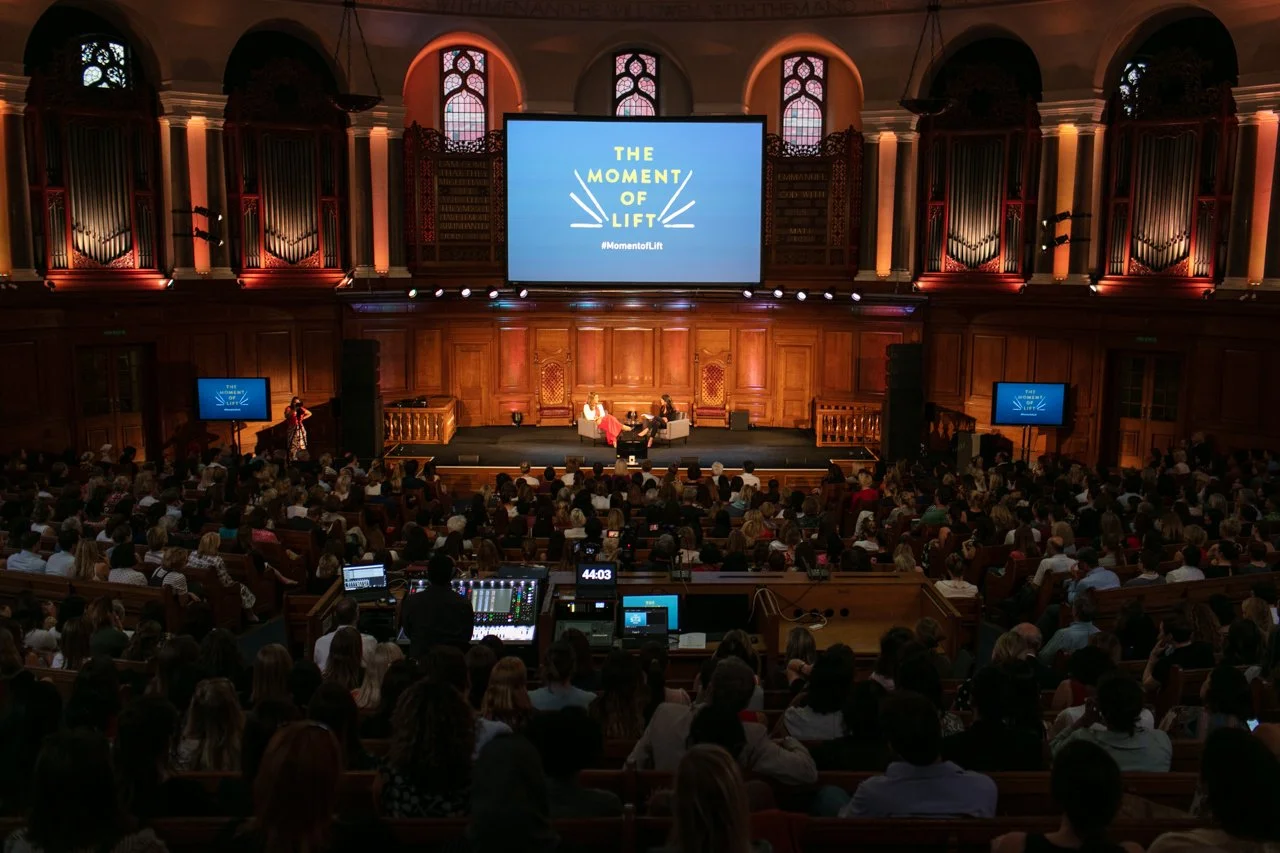 A large audience seated in a wooden auditorium with tall stained glass windows, watching a stage with two speakers and a large screen displaying 'The Moment of Lift' and the hashtag #MomentofLift.