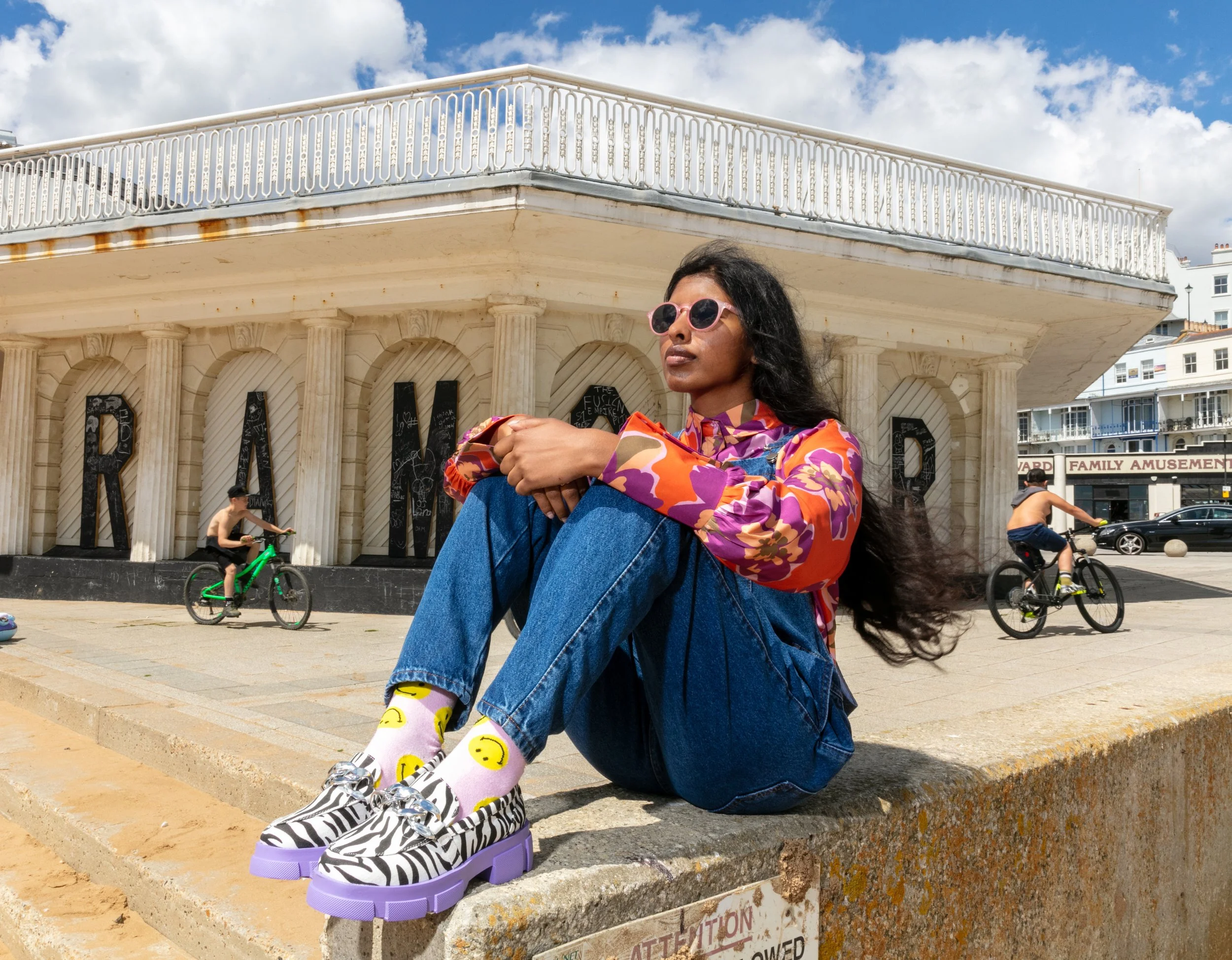 A woman with long dark hair wearing pink sunglasses, a colorful patterned shirt, and blue jeans sitting on a curb. She is wearing eye-catching platform shoes with a zebra stripe pattern and purple soles. In the background, there are people riding bik