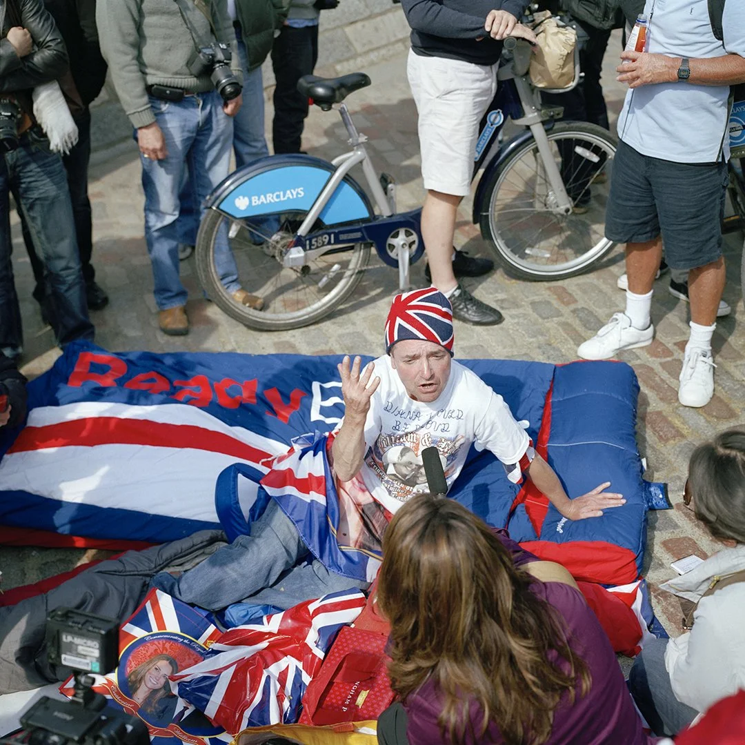 A man lying on a sleeping bag with British flags, surrounded by people. He is wearing a beanie with the Union Jack design, and appears to be speaking or explaining something, holding a microphone. Some people are standing nearby, with bicycles and ca