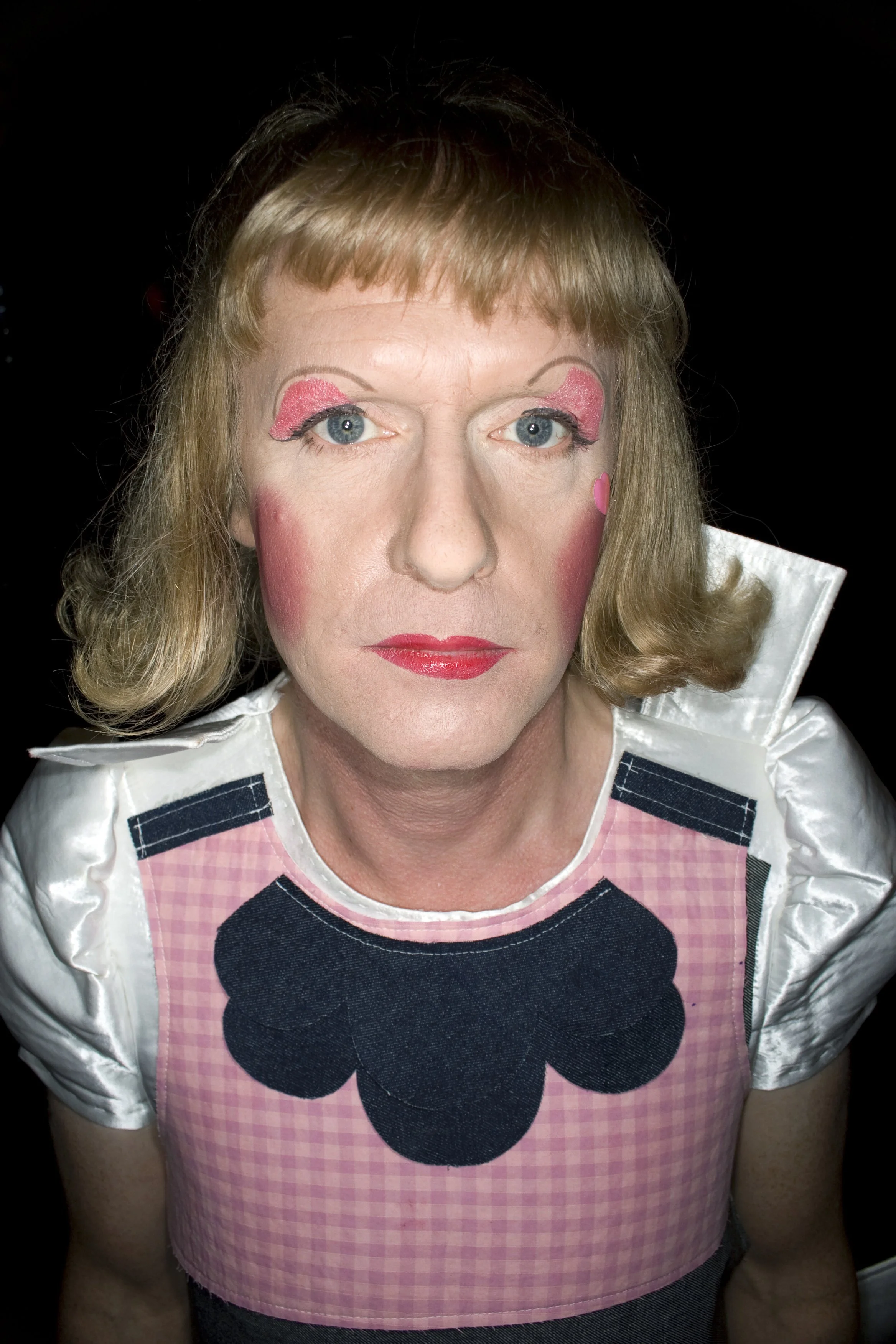 A person dressed as a clown with blue eyes, theatrical makeup, red lips, and blue and pink eyeshadow, wearing a dress with black flower appliqué and a white collar, against a black background.