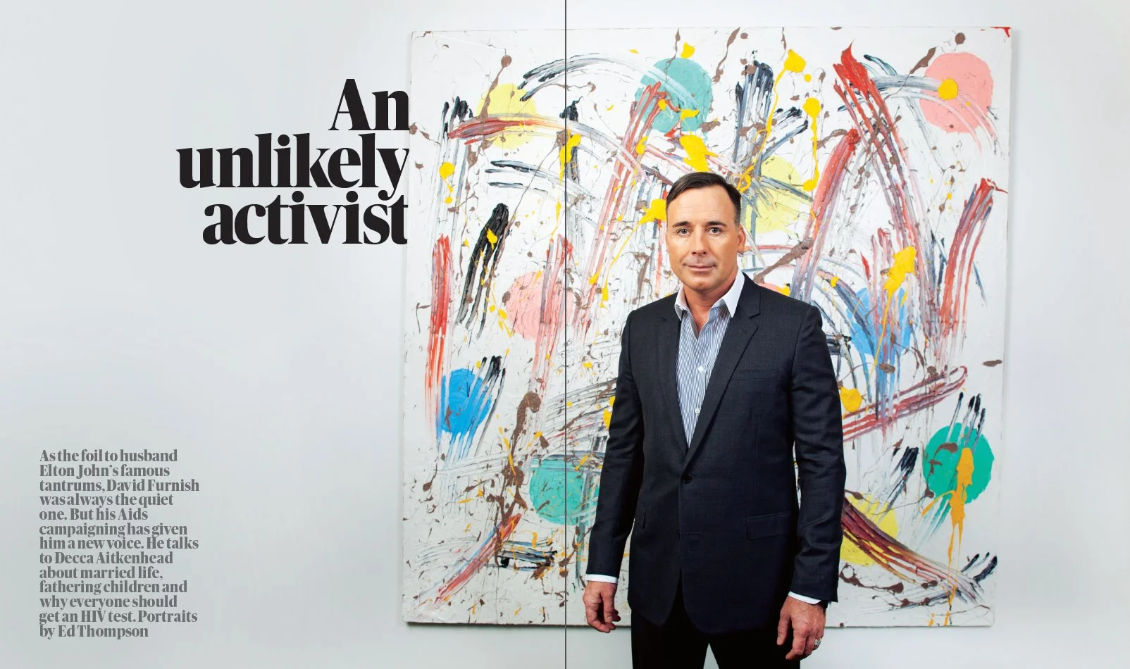 A man in a dark suit standing in front of an abstract colorful painting on a gallery wall.