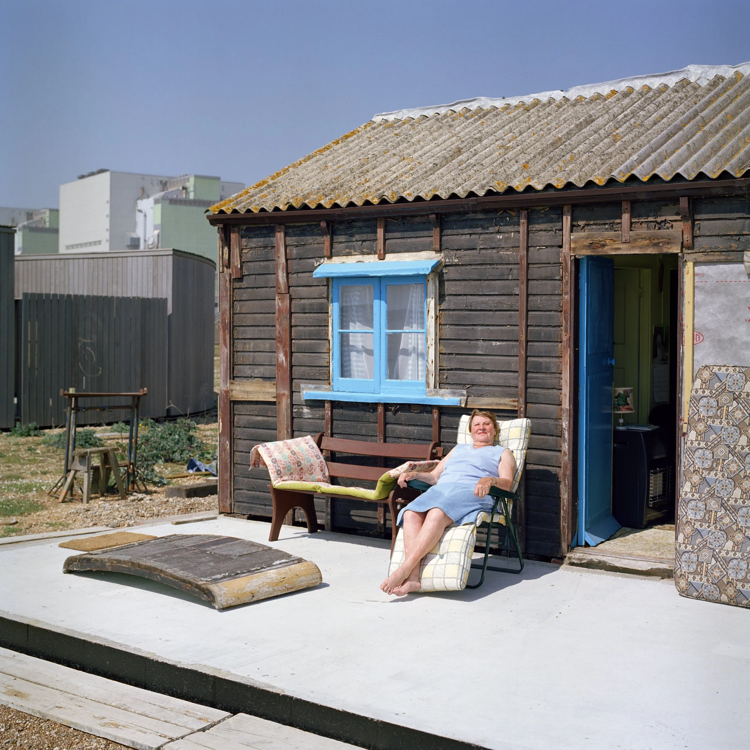 A woman relaxing outdoors on a deck chair in front of a rustic wooden building with a corrugated roof, next to a patterned pillow, with a porch light visible. There are modern buildings in the background and a clear blue sky overhead.