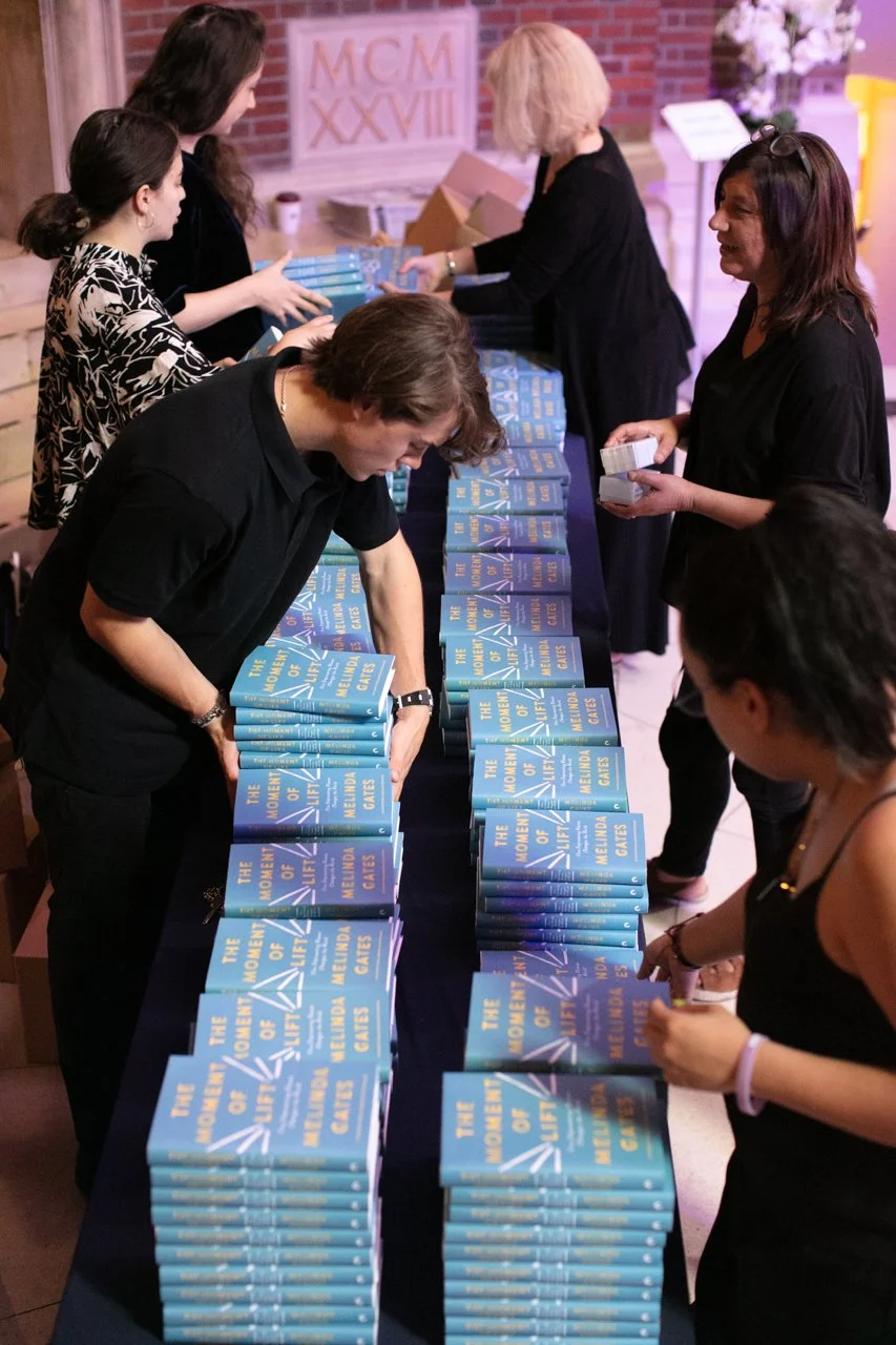 People at a book signing event with stacks of the book titled "The Moment of Life: The Woman's Guide to Melinda Cates" on a table.