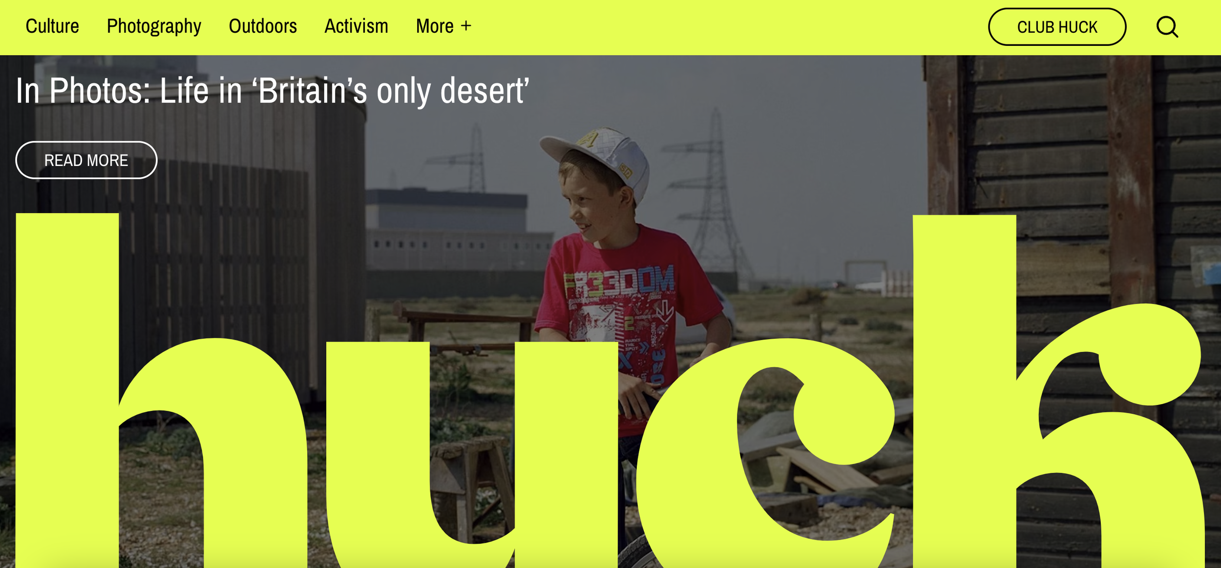Young boy wearing a white cap and red T-shirt with graphic design, standing outdoors in a rural or construction area with wooden structures, electrical towers, and industrial buildings in the background.