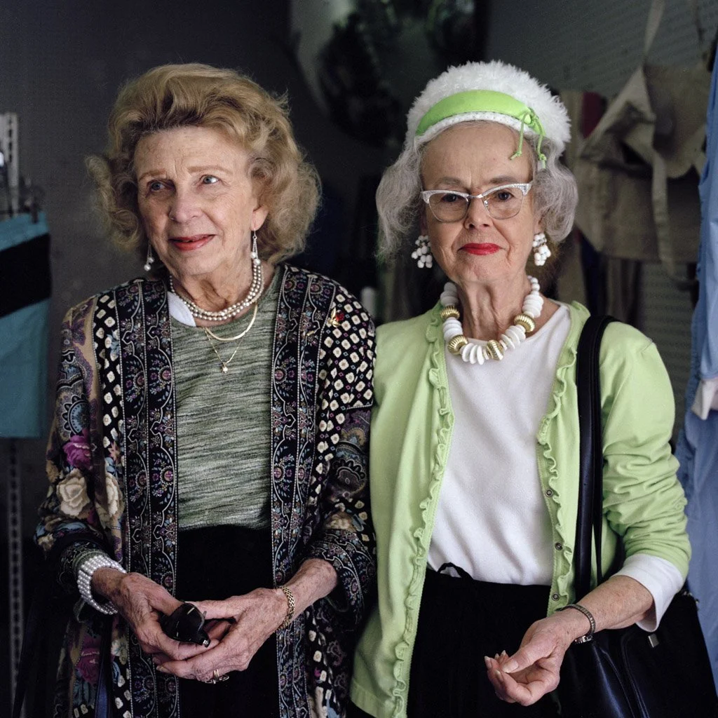 Two elderly women standing side by side; one is smiling and holding a small object, the other has a serious expression, wearing glasses and a bright green cardigan with accessories.