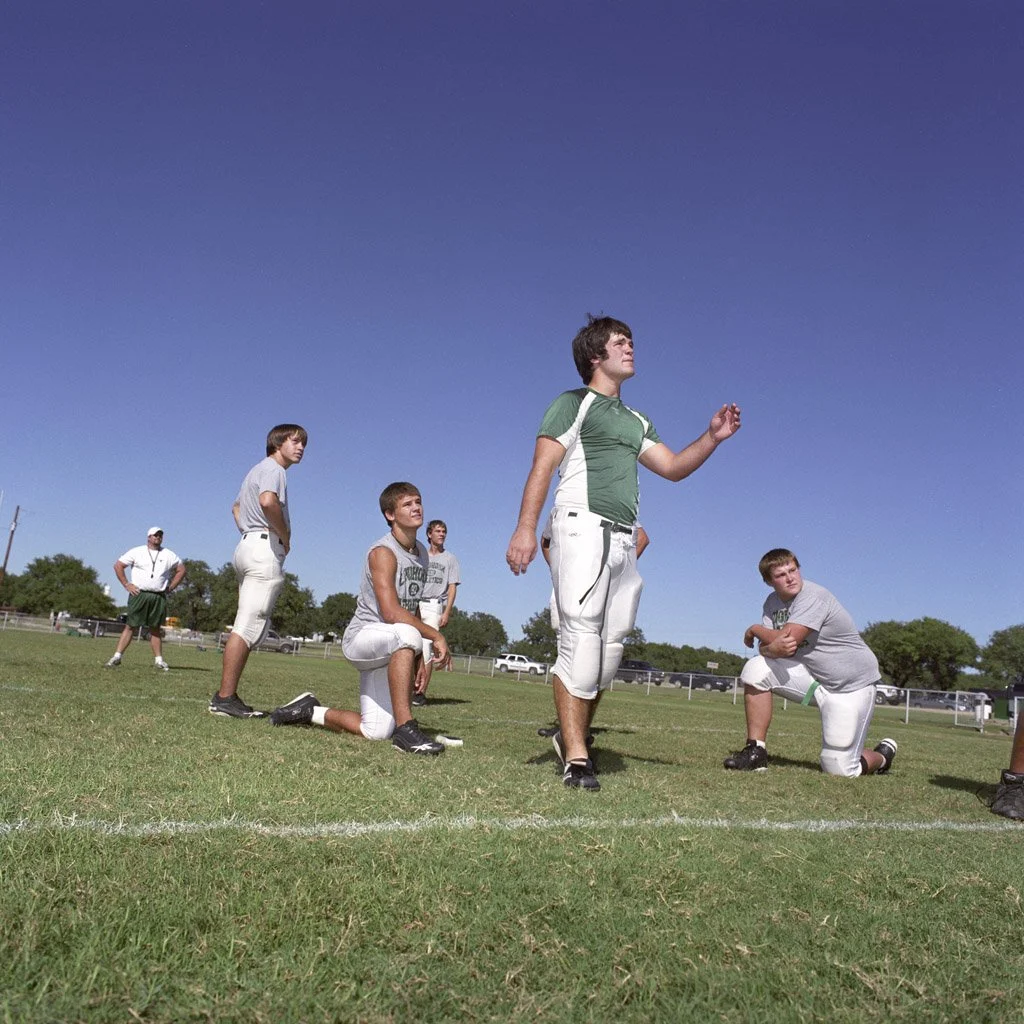 Group of young football players practicing on a field, with blue sky overhead, some kneeling and others standing in various positions.