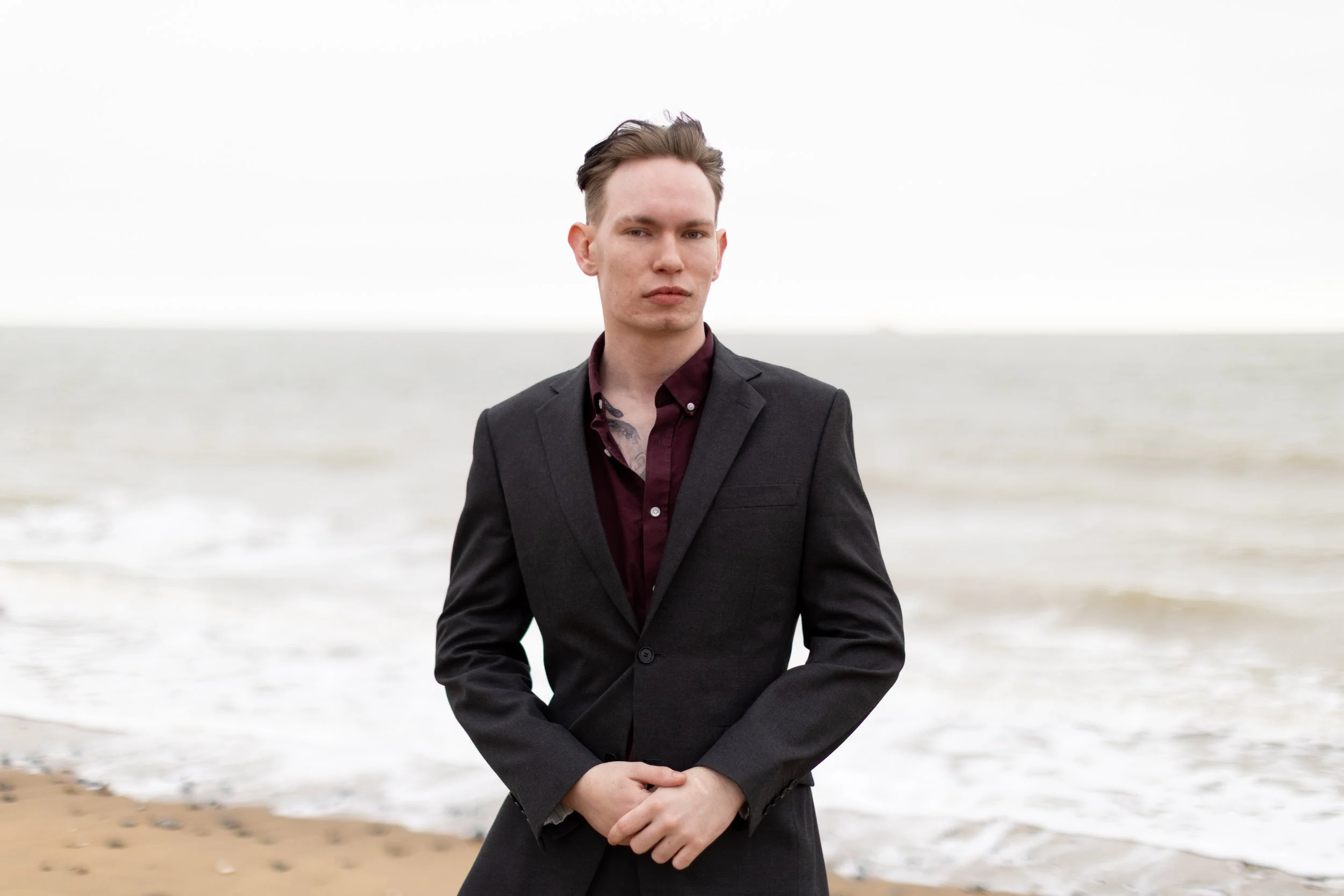A young man with short hair standing on a beach during daytime, wearing a dark blazer over a maroon shirt, with hands folded in front of him, facing slightly to the left with a serious expression.