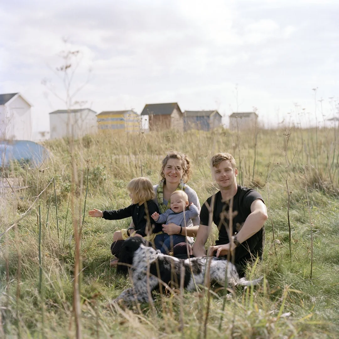 A family of four with two young children and a dog sitting on the grass in a field with small houses in the background on a sunny day.