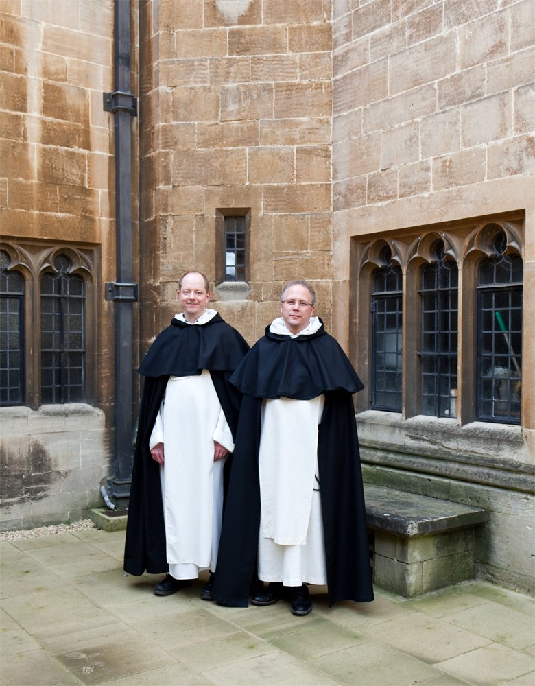 Two men standing outside near a stone building, dressed in traditional black and white academic robes with capes.