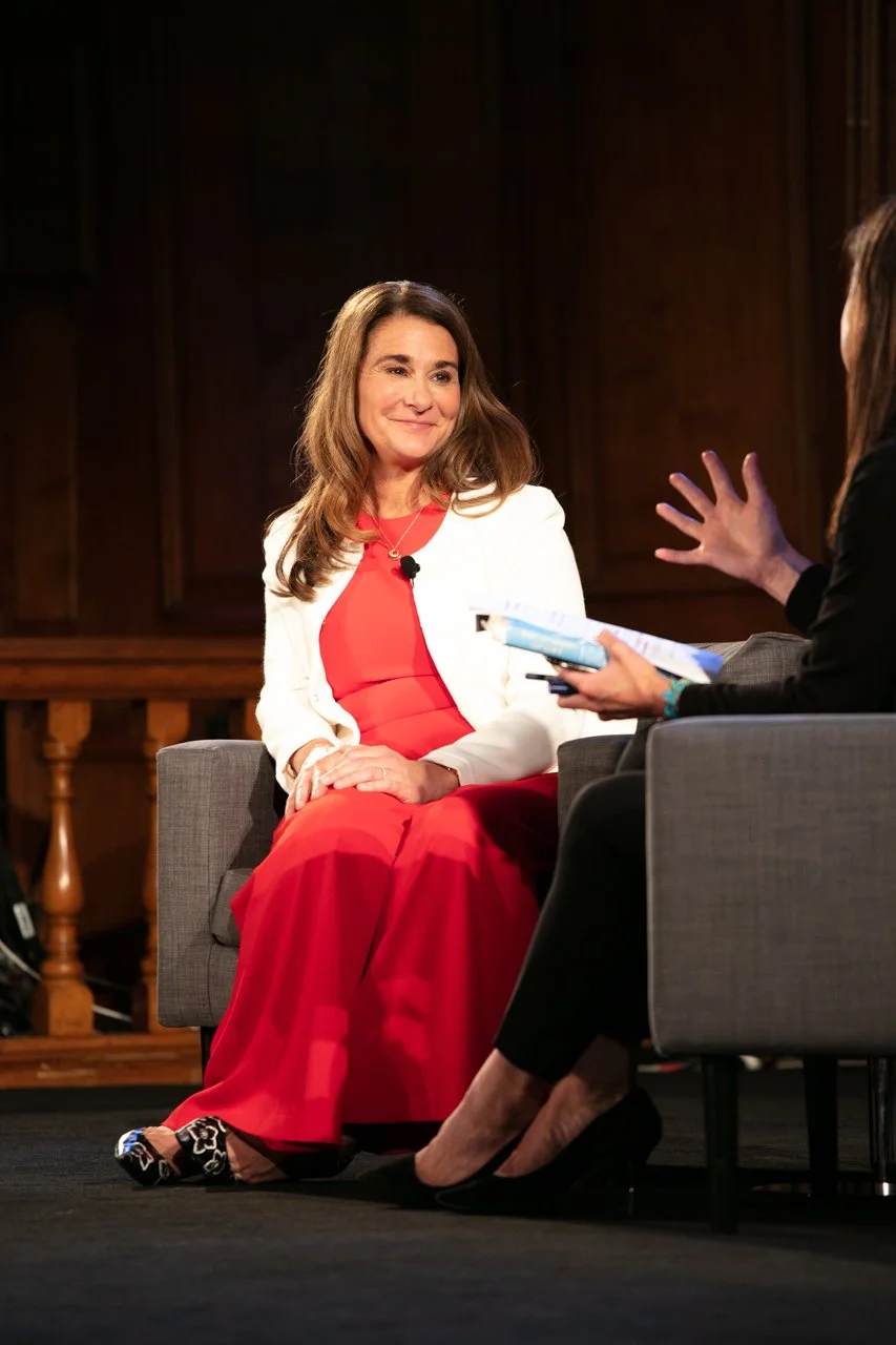 A woman in a white blazer and red dress sitting on a gray armchair, engaged in a conversation with another woman holding papers, in a wood-paneled room.