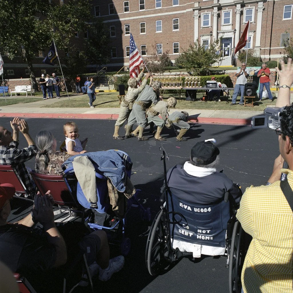Group of veterans performing a reenactment of the raising of the flag on the pavement in front of an audience, including children and elderly people, with some waving flags and clapping, outside a large brick building.