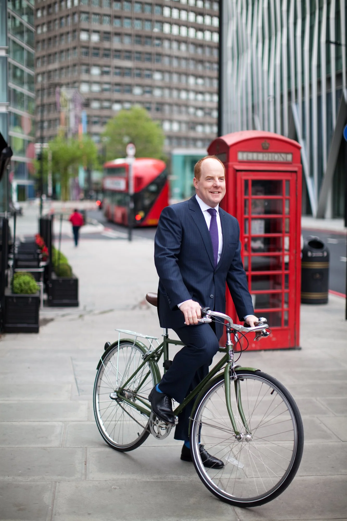 A man in a suit riding a bicycle on a city sidewalk with a red telephone booth and a double-decker bus in the background.