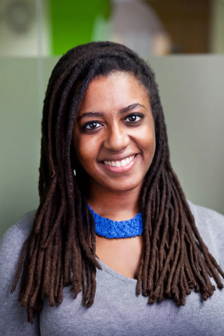 A smiling woman with long dreadlocks, wearing a blue necklace and a gray top, standing in front of a blurred background with green and gray.